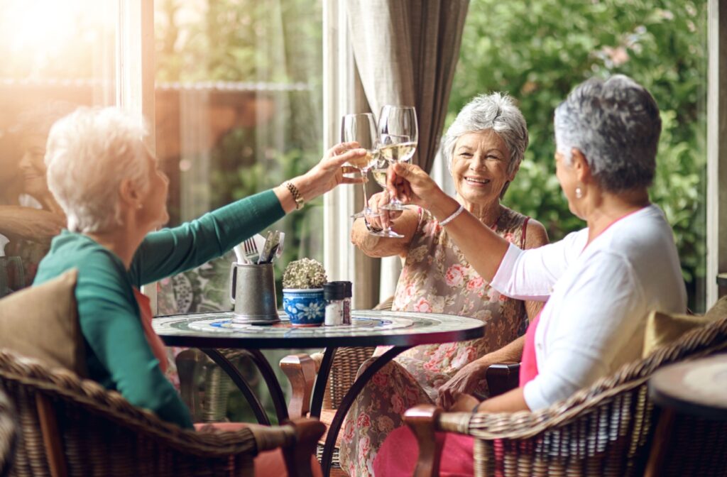 Three senior women sit around a small table on a patio, raising their glasses in a toast.